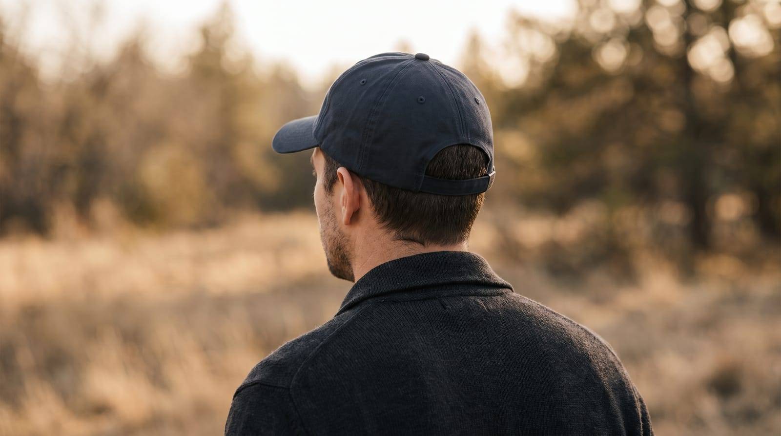 A man in a plain navy baseball cap photographed from behind in soft outdoor daylight, looking away across open ground, the everyday habit blamed for hair loss more often than any other